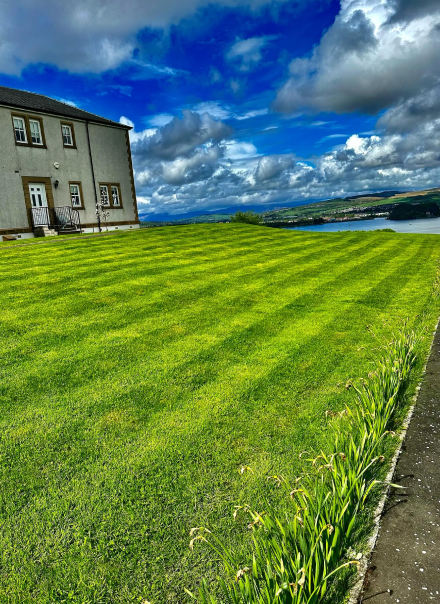 A beautifully mowed grass lawn with stripes in an area that looks like the north of Scotland with an old fashioned cottage building on a hillock on a beautiful summers day with the North Sea in the background